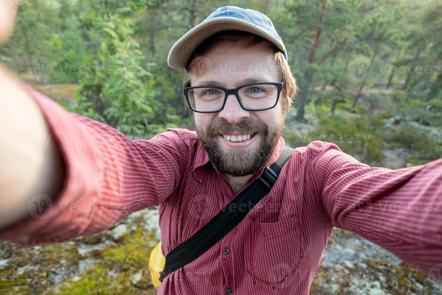 Ein glücklicher, lächelnder Mann mit Mütze und Brille macht ein Selfie auf dem Hintergrund des Waldes. Freizeitaktivitäten. foto