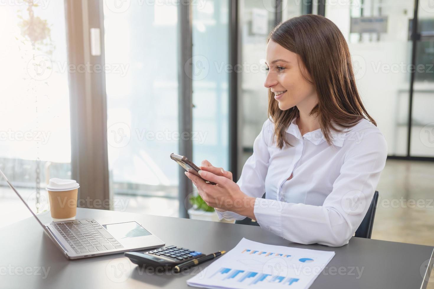 marketing, finanzen, buchhaltung, planung, weiße biracial geschäftsfrau, die mit dem handy kunden kontaktiert und sie über geschäftspartnerschaften des unternehmens auf dem bürotisch mit laptop und dokumenten informiert. foto