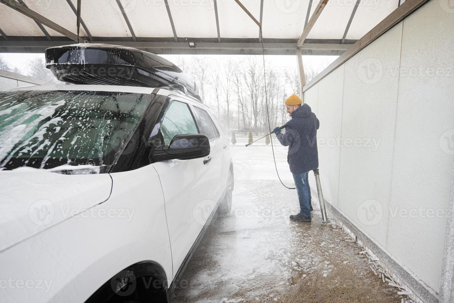 Mann wäscht Hochdruckwasser amerikanisches SUV-Auto mit Dachgepäckträger bei Selbstbedienungswäsche bei kaltem Wetter. foto