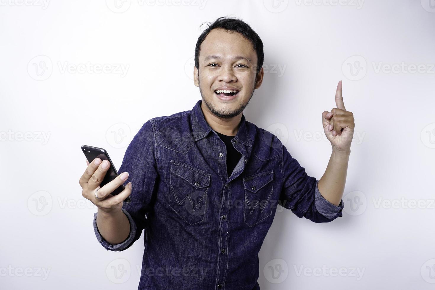 ein Porträt eines glücklichen asiatischen Mannes, der ein blaues Hemd trägt und ihr Telefon hält, isoliert durch weißen Hintergrund foto