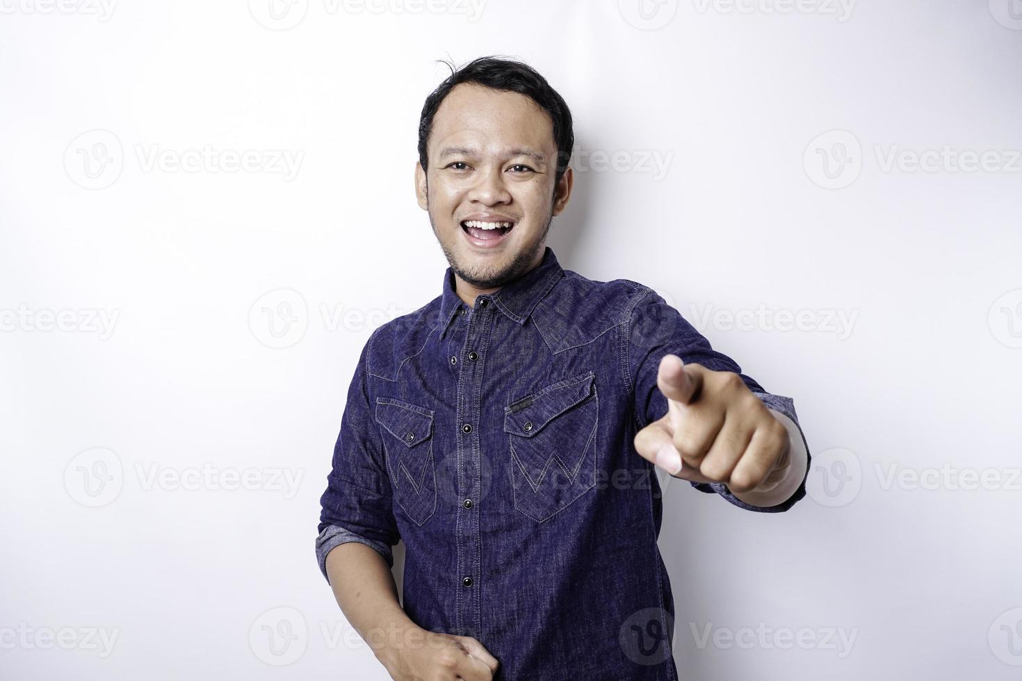 Junger asiatischer Mann mit blauem Hemd, der vor isoliertem weißem Hintergrund steht und mit glücklichem Gesicht mit dem Finger auf die Kamera zeigt. gute Energie und Stimmung. foto