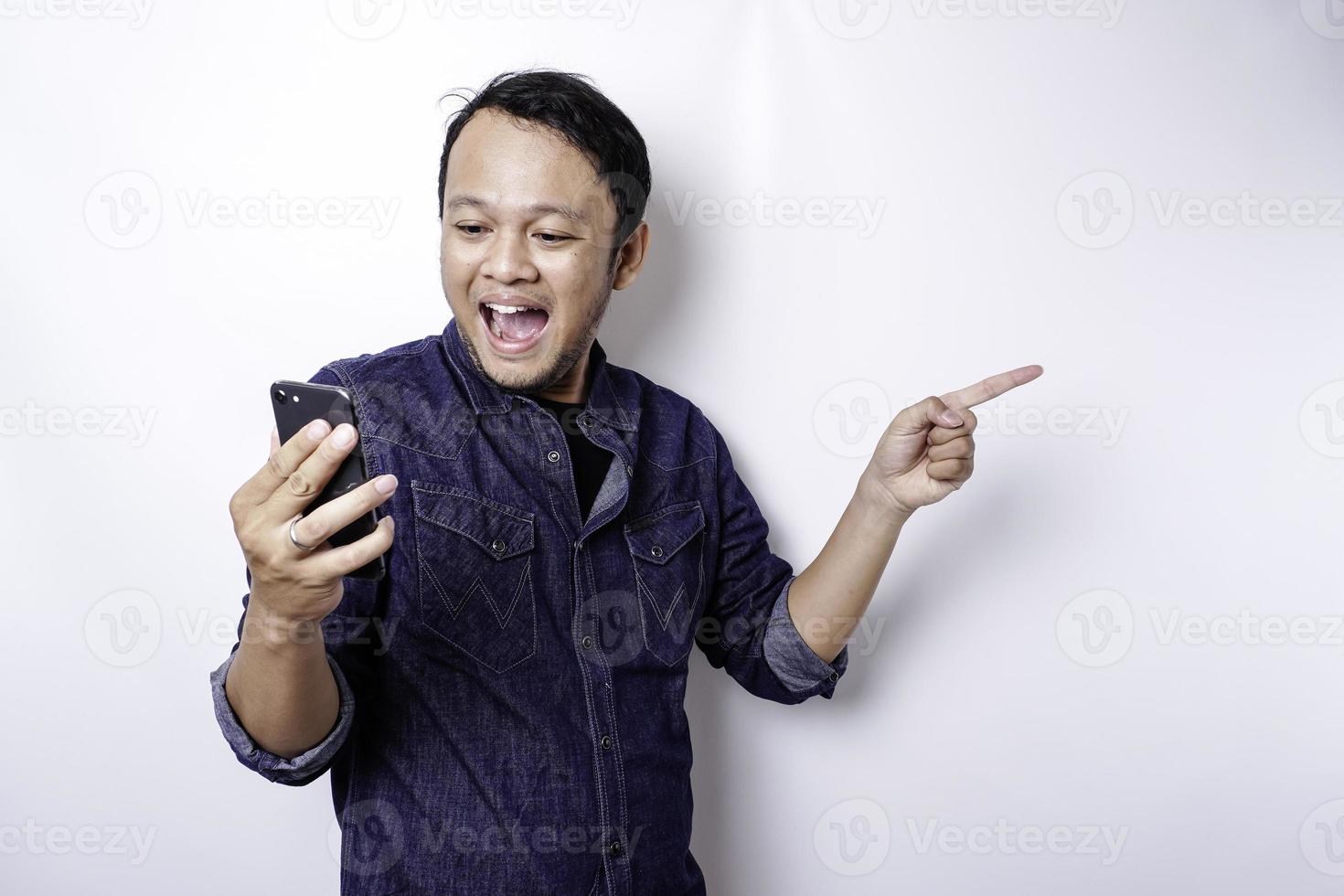 Aufgeregter asiatischer Mann mit blauem Hemd, der auf den Kopierbereich neben ihm zeigt, während er sein Telefon hält, isoliert durch weißen Hintergrund foto