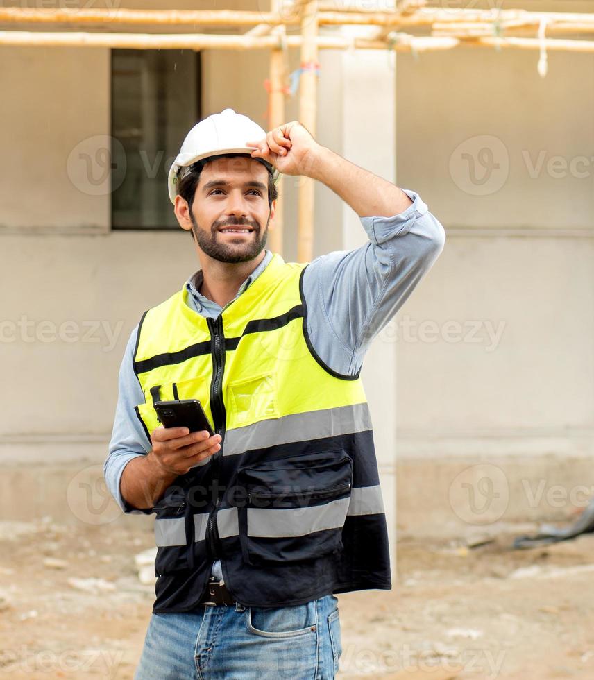 Männlicher Architekt oder Ingenieur, der während der Arbeit auf der Baustelle ein Mobiltelefon zur Kommunikation verwendet. professioneller bauunternehmer, der auf dem standort der wohnsiedlung steht, nutzt smartphone bei der arbeit. foto