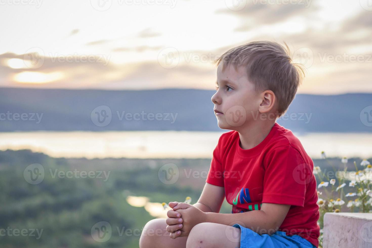 Süßer Junge in einem roten T-Shirt auf dem Hintergrund eines atemberaubenden Sonnenuntergangs. Reise. Das Gesicht drückt natürliche freudige Emotionen aus. nicht inszenierte Fotos aus der Natur.