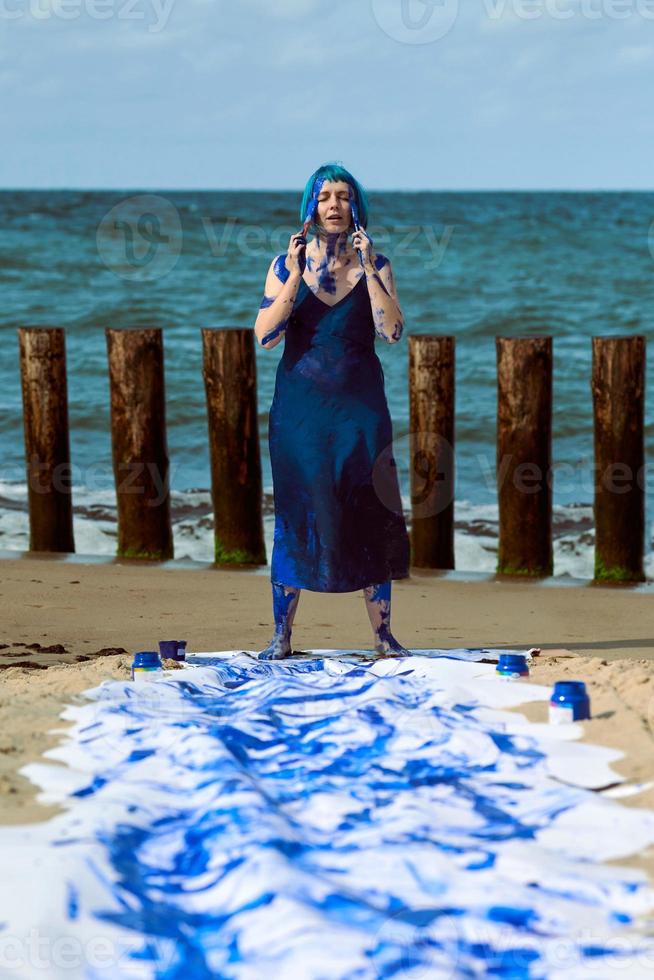 glückliche Performance-Künstlerin in blauem Kleid, beschmiert mit blauen Gouachefarben, tanzt am Strand foto