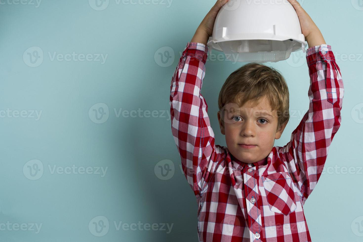 Ein süßer Junge in einem karierten Hemd hält einen Bauhelm über dem Kopf, ein Porträt eines kleinen Baumeisters foto