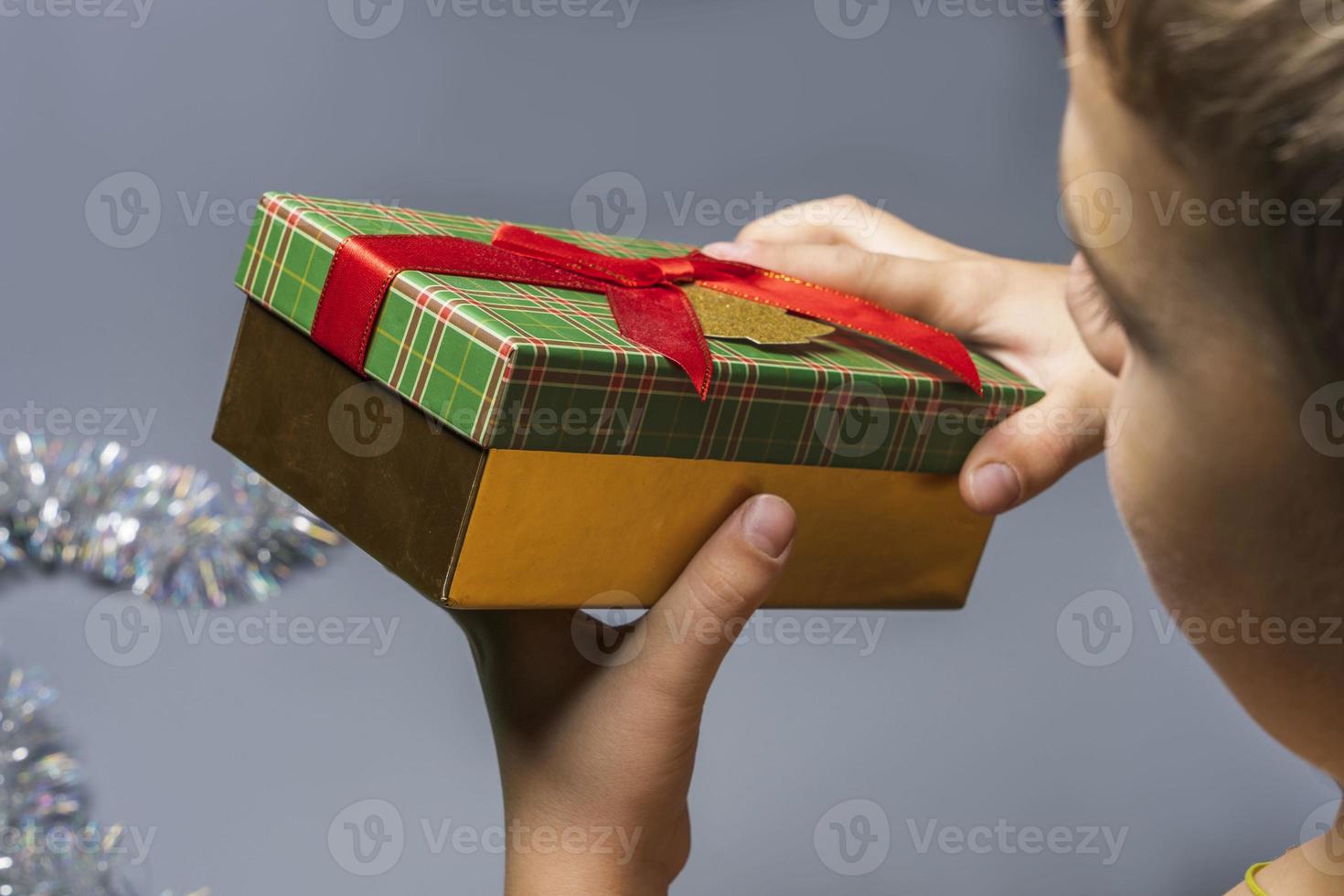 Ein Junge mit einem Weihnachtsgeschenk in den Händen auf grauem Hintergrund, eine Schachtel mit einem Neujahrsgeschenk in den Händen eines Kindes foto