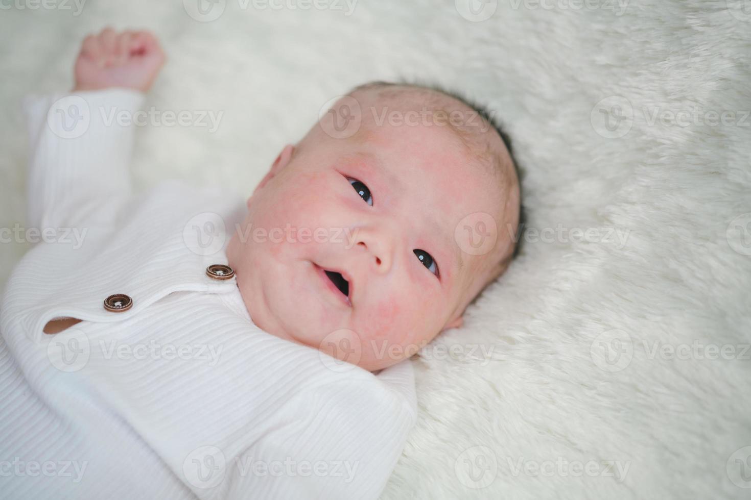 nahaufnahme süßes neugeborenes baby im weißen bodysuit, der sich allein auf dem bett hinlegt. entzückendes kind ruht auf weißen bettlaken und starrt friedlich in die kamera. Säuglings-, Gesundheitswesen und Pädiatrie, Säuglingskonzept foto