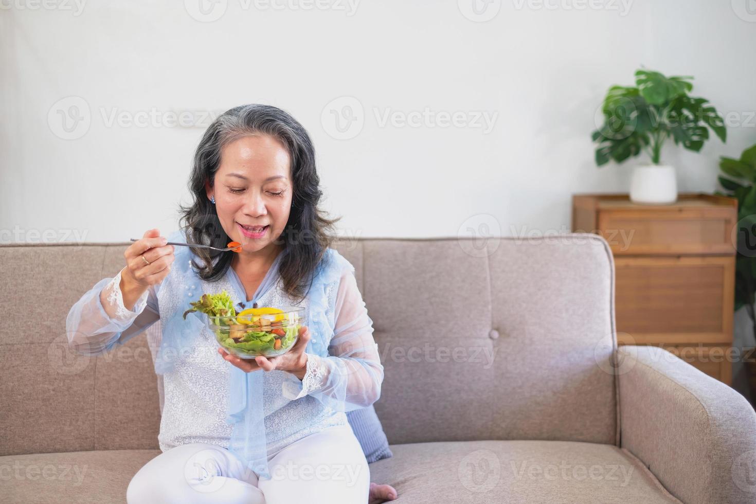 asiatische Seniorin, die Gemüsesalat und gesundes Essen isst und glücklich auf dem Sofa im Haus für einen gesunden Körper isst. gesundes lebensmittelkonzept foto