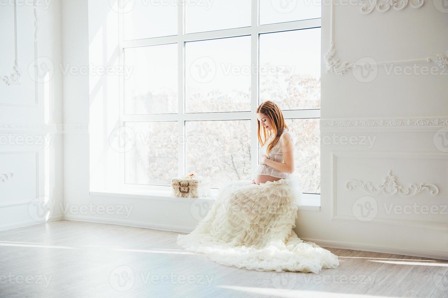 Rothaariges schwangeres junges Mädchen in einem weißen Kleid in der Nähe des Fensters foto