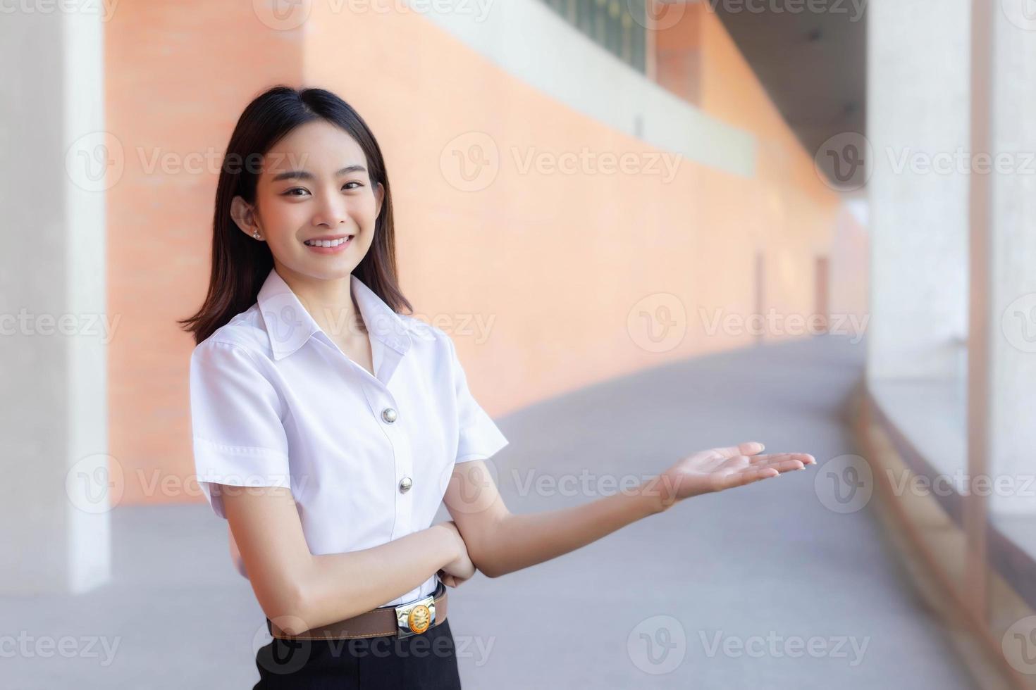 asiatische schöne junge studentin lächelt und schaut in die kamera, die steht, um etwas selbstbewusst im universitätshintergrund zu präsentieren foto