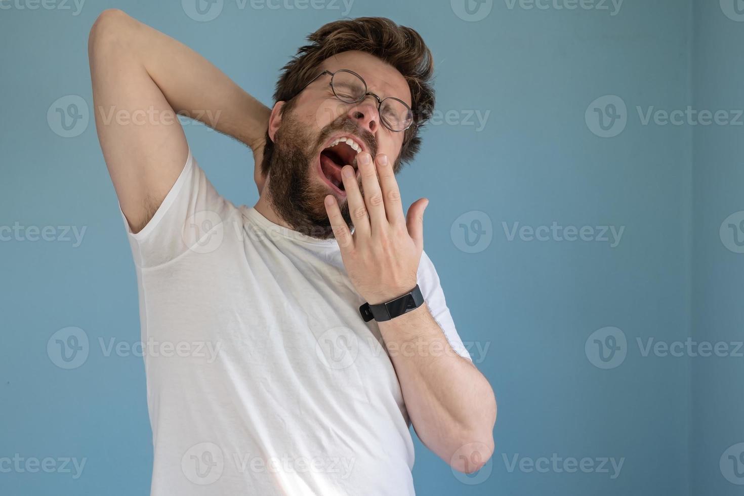 bärtiger mann mit brille und weißem t-shirt gähnt mit weit geöffnetem mund und bedeckt ihn mit der hand, er ist schläfrig oder gelangweilt. Blauer Hintergrund. foto