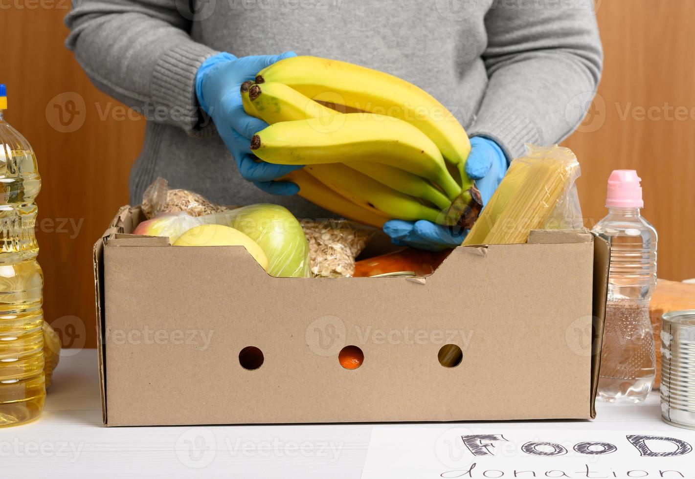 Frau in Handschuhen sammelt Lebensmittel, Obst und Dinge und einen Karton, um Bedürftigen zu helfen foto