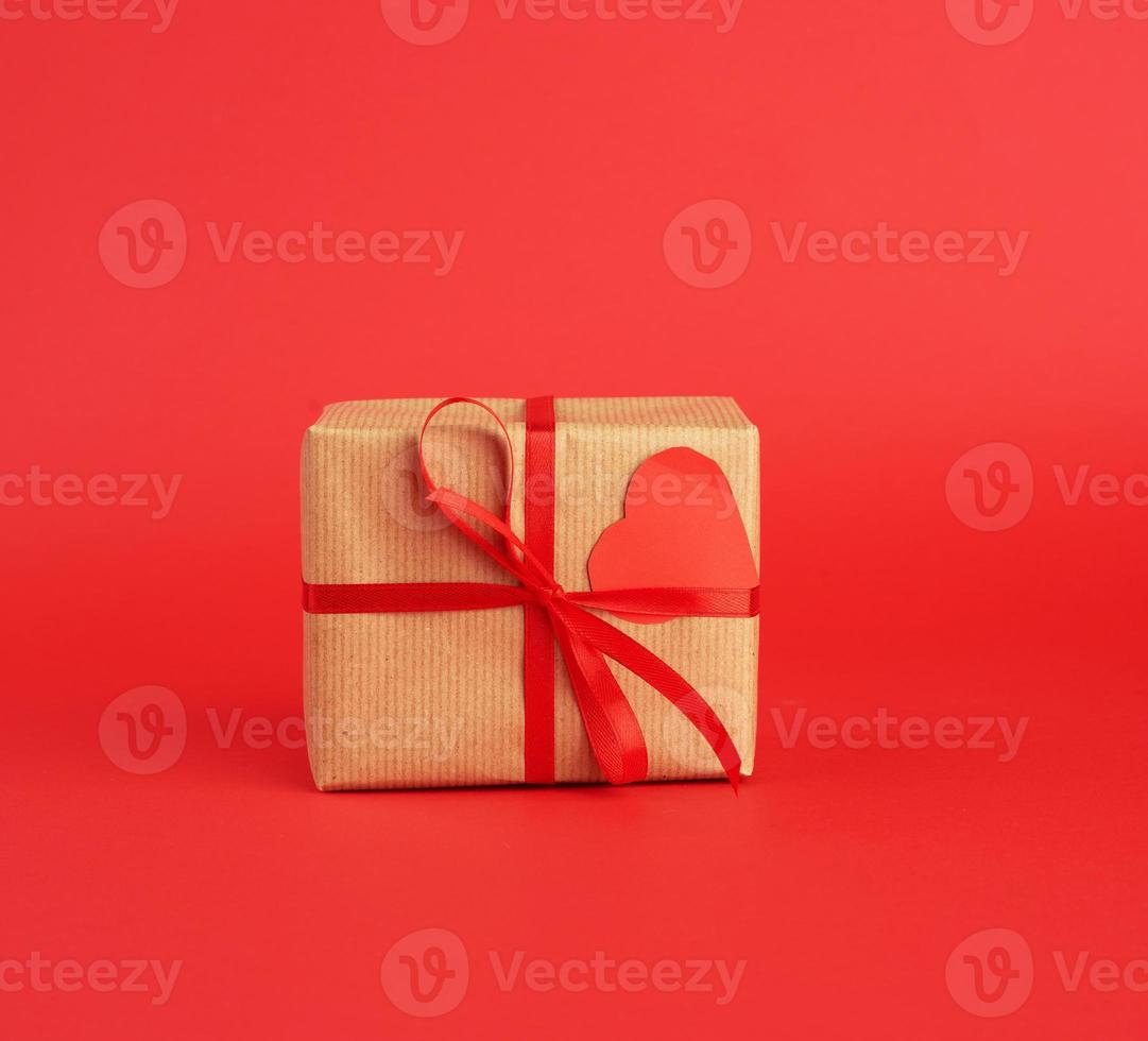 Eingewickelte quadratische Schachtel mit einem Geschenk in braunem Bastelpapier und mit einem dünnen roten Seidenband gebunden foto