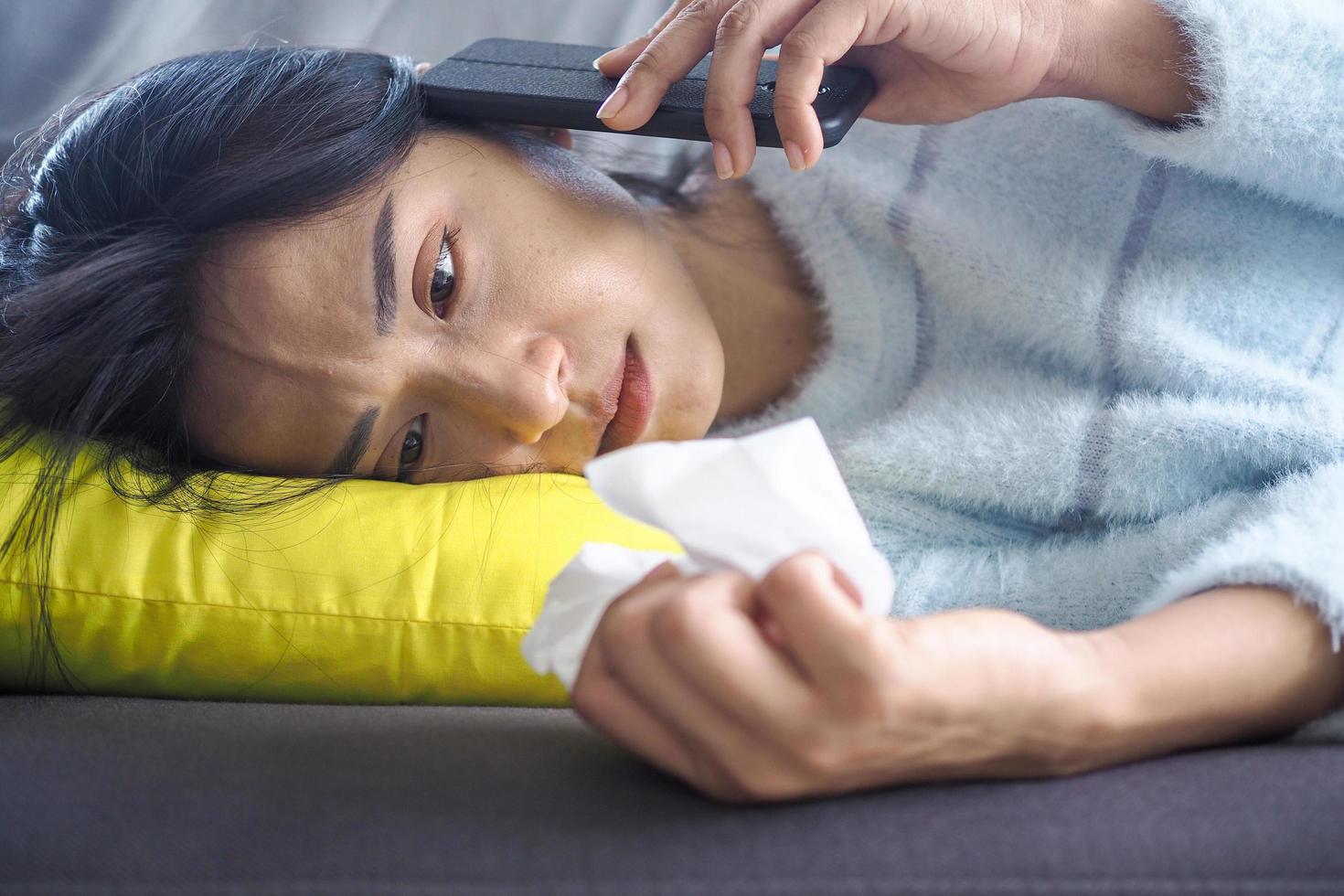 asiatische frau ist an erkältung und fieber erkrankt. auf dem sofa im haus liegen, mit dem telefon sprechen und ein trauriges gesicht haben. foto