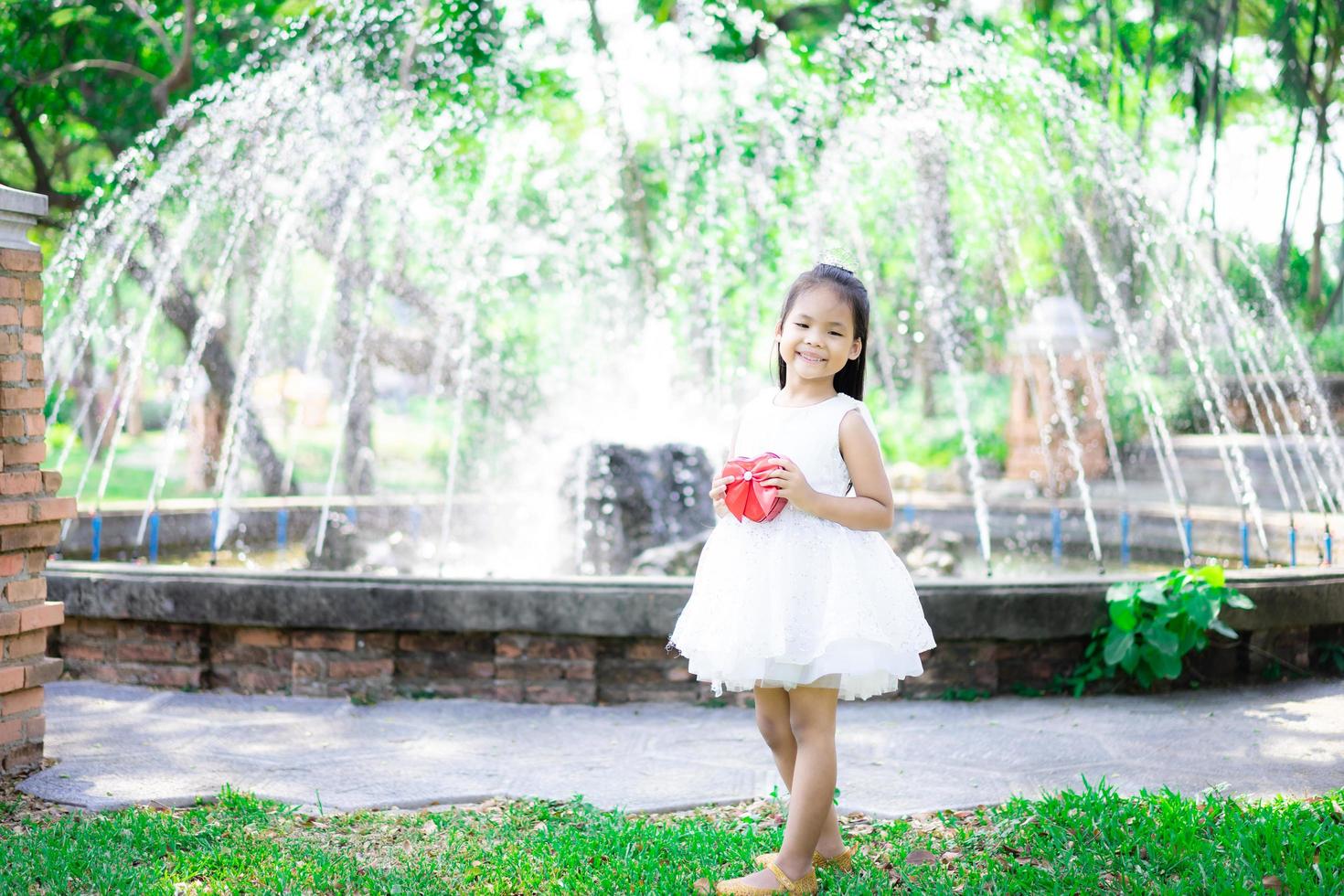 süßes kleines asiatisches Mädchen im weißen Kleid, das ein rotes Geschenk im Park hält foto