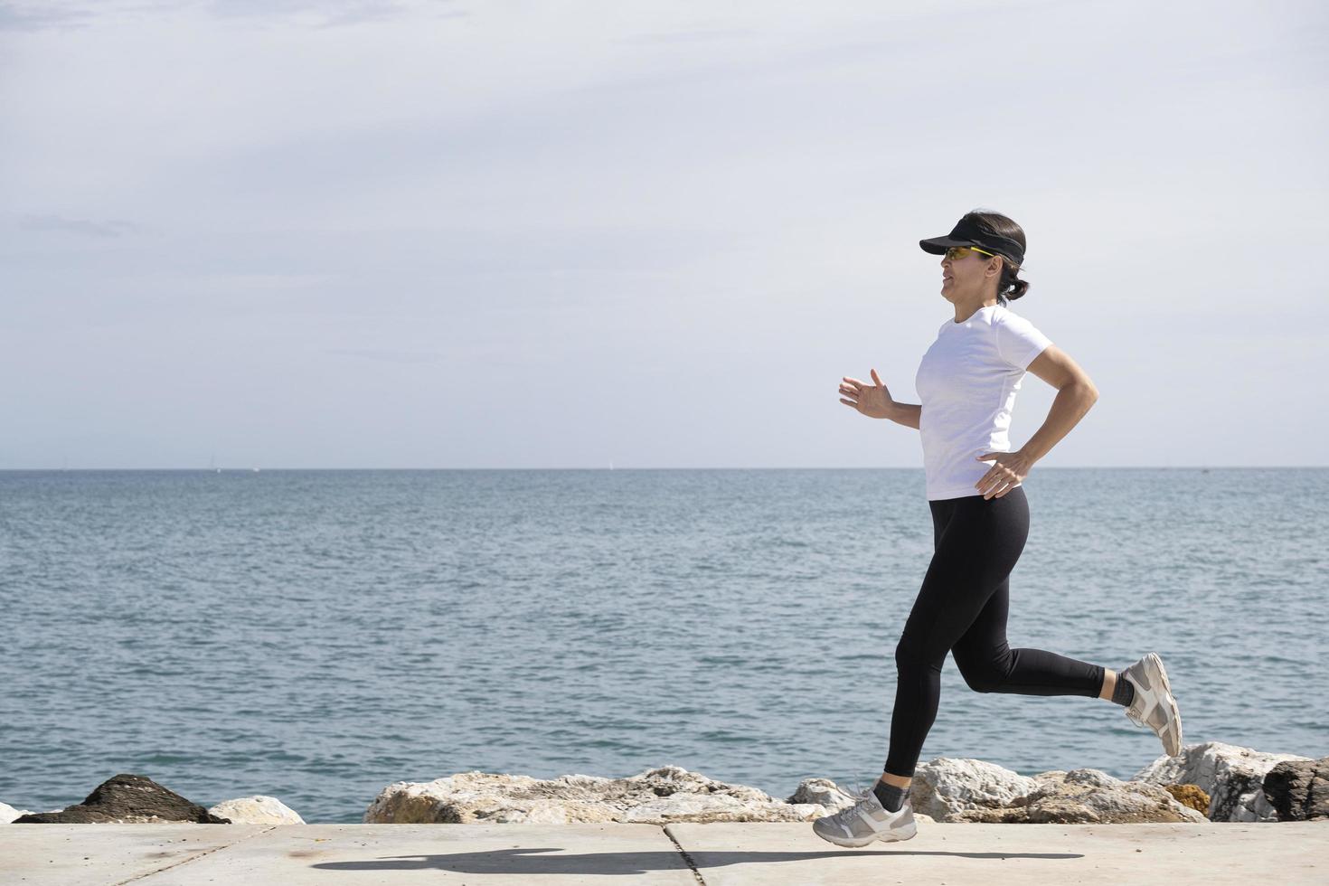 Frau mit Mütze und Sonnenbrille läuft entlang der Promenade, das Meer im Hintergrund foto