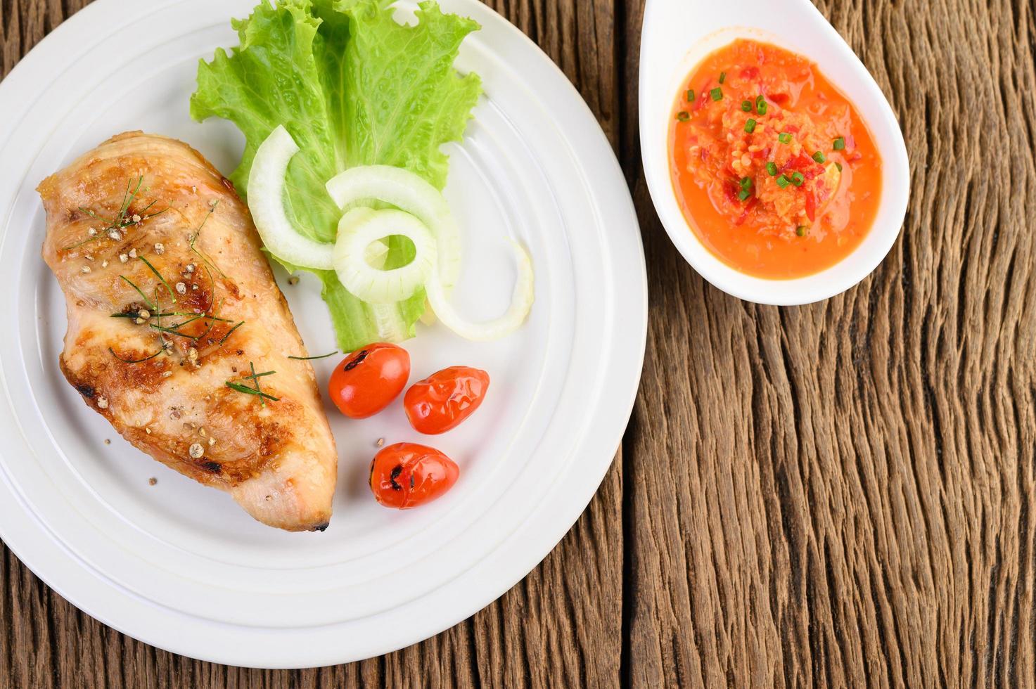 Gegrilltes Hähnchen auf einem Holztisch mit Tomaten, Salat, Zwiebeln und Chilisauce foto