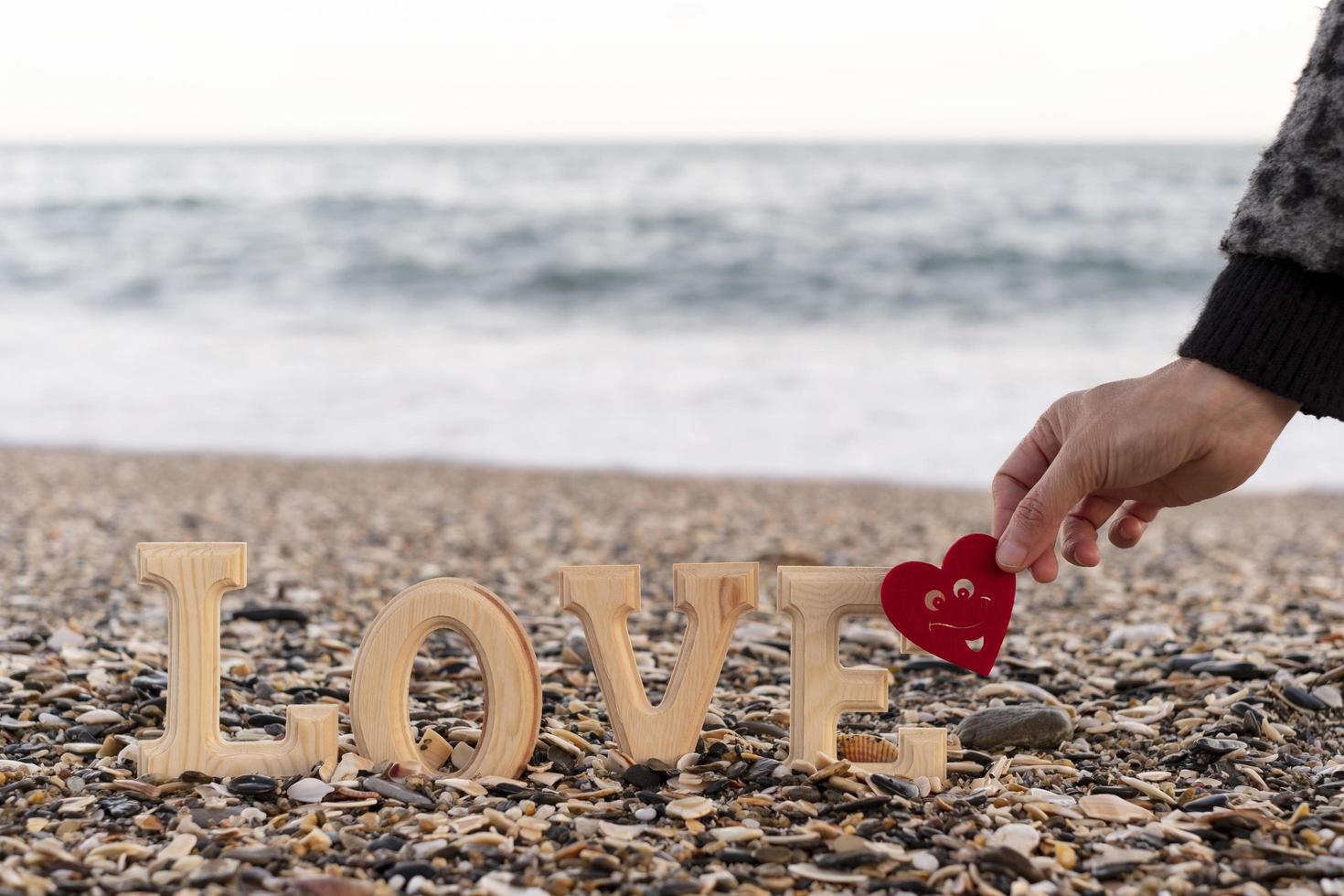 Holzbuchstaben, die das Wort Liebe und eine Hand bilden, die ein rotes Herz an der Küste hält. Konzept von st. Valentinstag foto