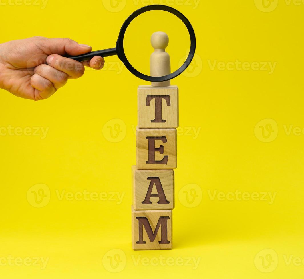 Holzfigur eines Mannes auf einem Sockel und einer Hand mit einer Lupe auf gelbem Hintergrund. das Konzept der Rekrutierung und Suche nach talentierten Mitarbeitern foto