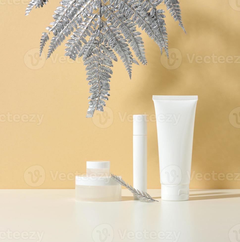 weiße tube für kosmetik, ein glas creme und blattsilber auf einem weißen tisch. Kosmetik auf einem beigen Schattenhintergrund. cremeflasche, lotion, reiniger, shampoo für die hautpflege foto