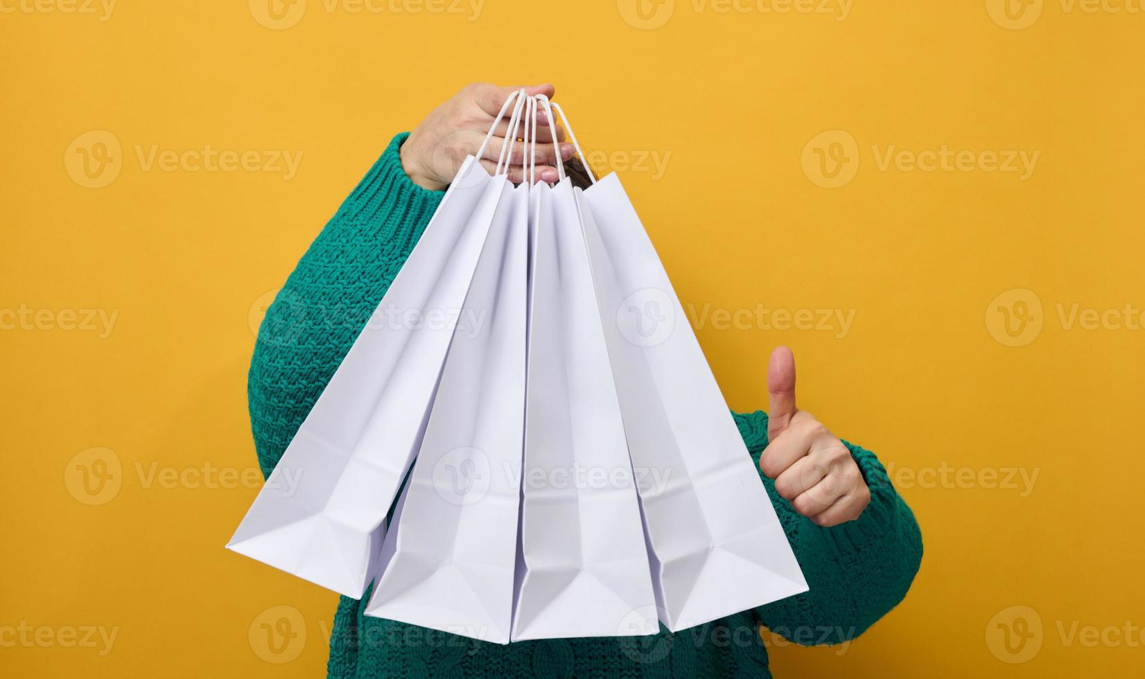 eine frau in einem pullover hält weiße papiertüten auf gelbem hintergrund und zeigt eine ähnliche geste mit der hand. Einkäufe. saisonaler Verkauf foto