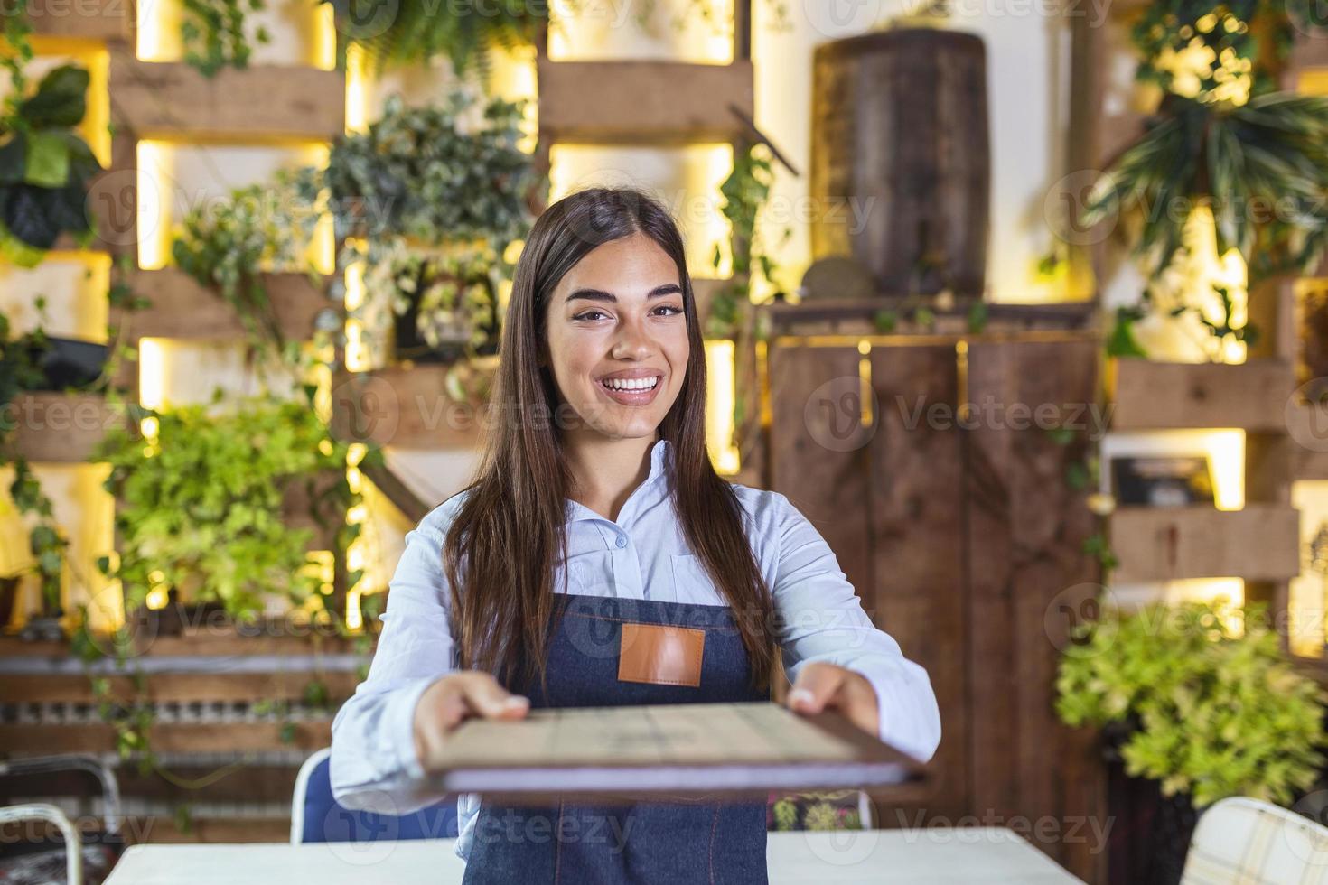 Fröhliche, schöne, lächelnde Kellnerin mit Schürze, die in einem Restaurant ein Ordnermenü gibt, in die Kamera schaut, in einem gemütlichen Kaffeehaus steht, guter Service foto