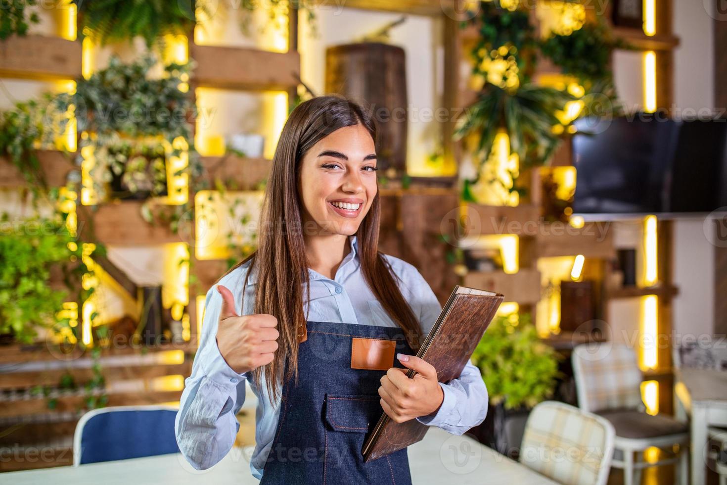 Fröhliche, schöne, lächelnde Kellnerin mit Schürze, die in einem Restaurant ein Ordnermenü gibt, in die Kamera schaut, in einem gemütlichen Kaffeehaus steht, guter Service foto