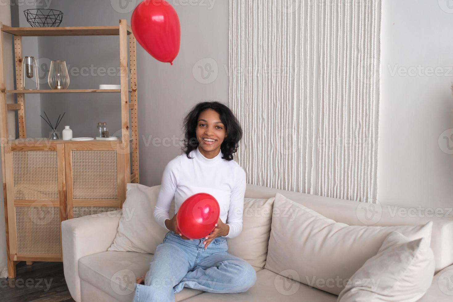 Afroamerikanisches Mädchen, das auf dem Sofa mit roten Luftballons in Herzform spielt. Valentinstag. foto