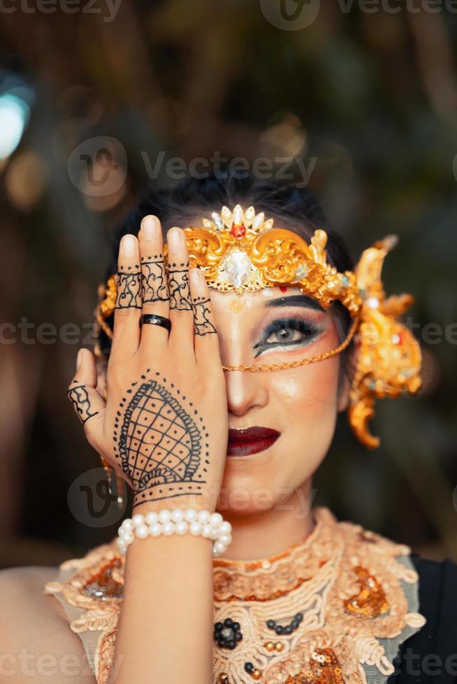 asiatische Frau mit Make-up, die sein Gesicht mit ihrer Hand bedeckt, während sie geschminkt ist und eine goldene Krone und eine goldene Tiara trägt foto