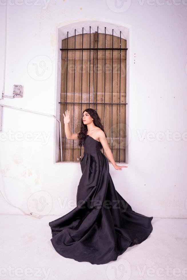 traurige asiatin, die vor dem braunen holzfenster steht, während sie ein schwarzes kleid trägt foto