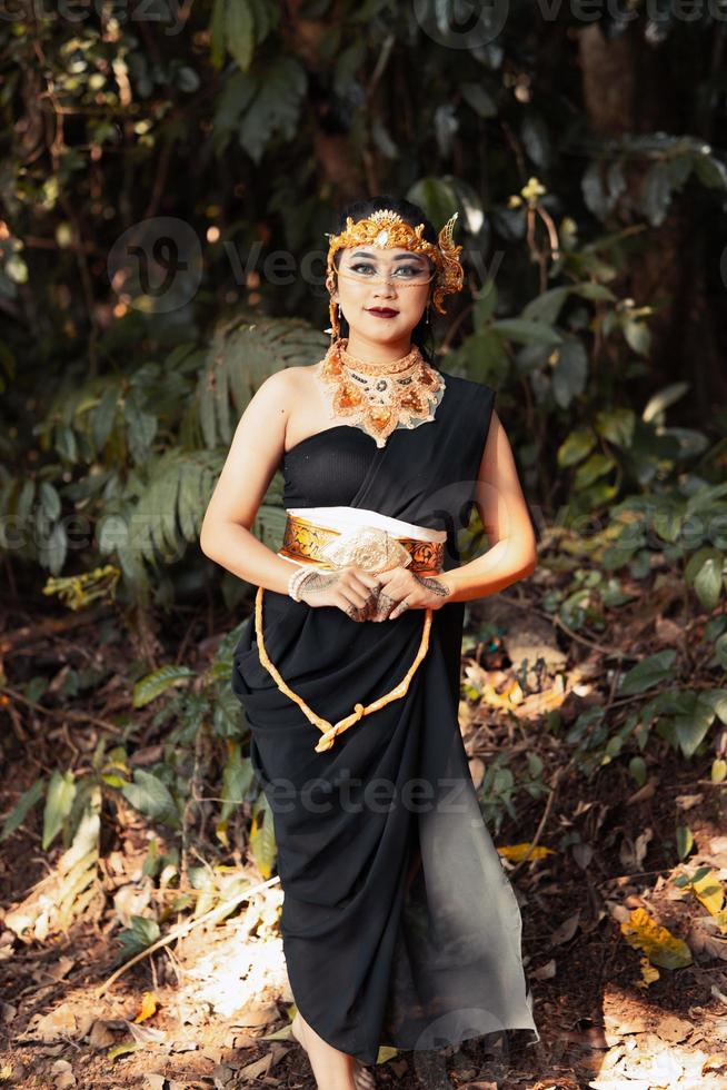 Schöne asiatische Frau, die in einem schwarzen Kleid steht, während sie eine goldene Krone und eine goldene Kette im Wald trägt foto