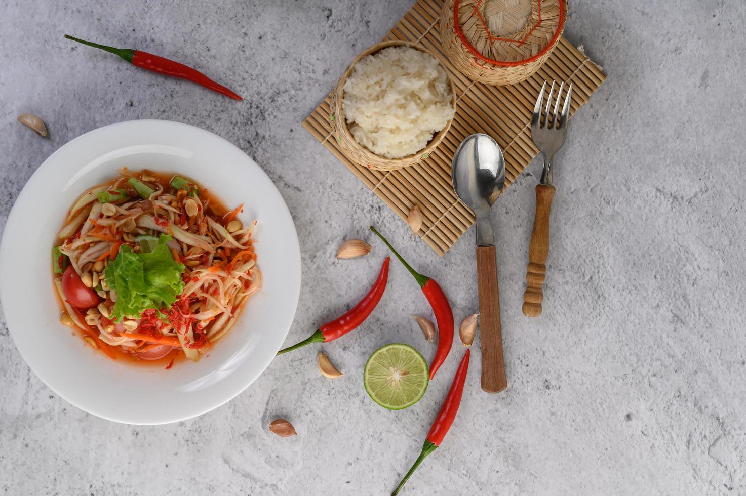thailändischer Papayasalat auf einem weißen Teller mit Klebreis, Chili, Löffel und Gabel foto