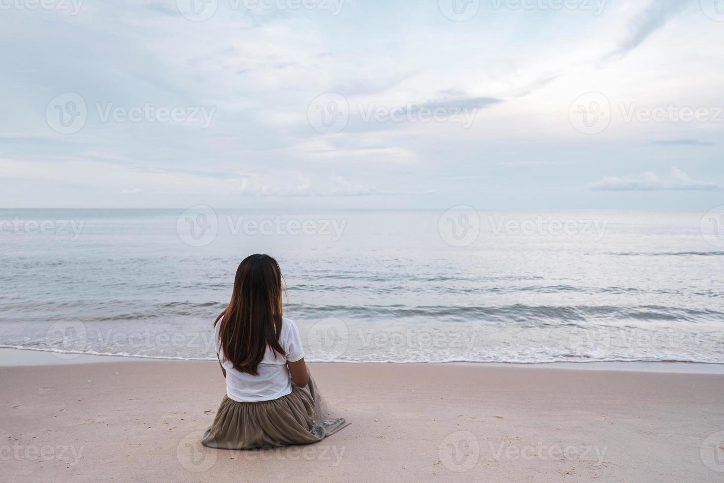 Einsame junge Asiatin, die bei Sonnenuntergang am Strand sitzt foto