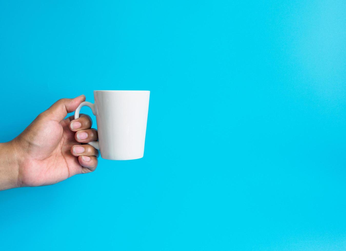 mannhand, die eine weiße kaffeetasse hält, gefüllt mit heißem schwarzem kaffee americano, kein zucker, keine milch, trinkfertig, erfrischend. aroma wach frisch zur arbeit platziert auf einem blau isolierten hintergrund foto