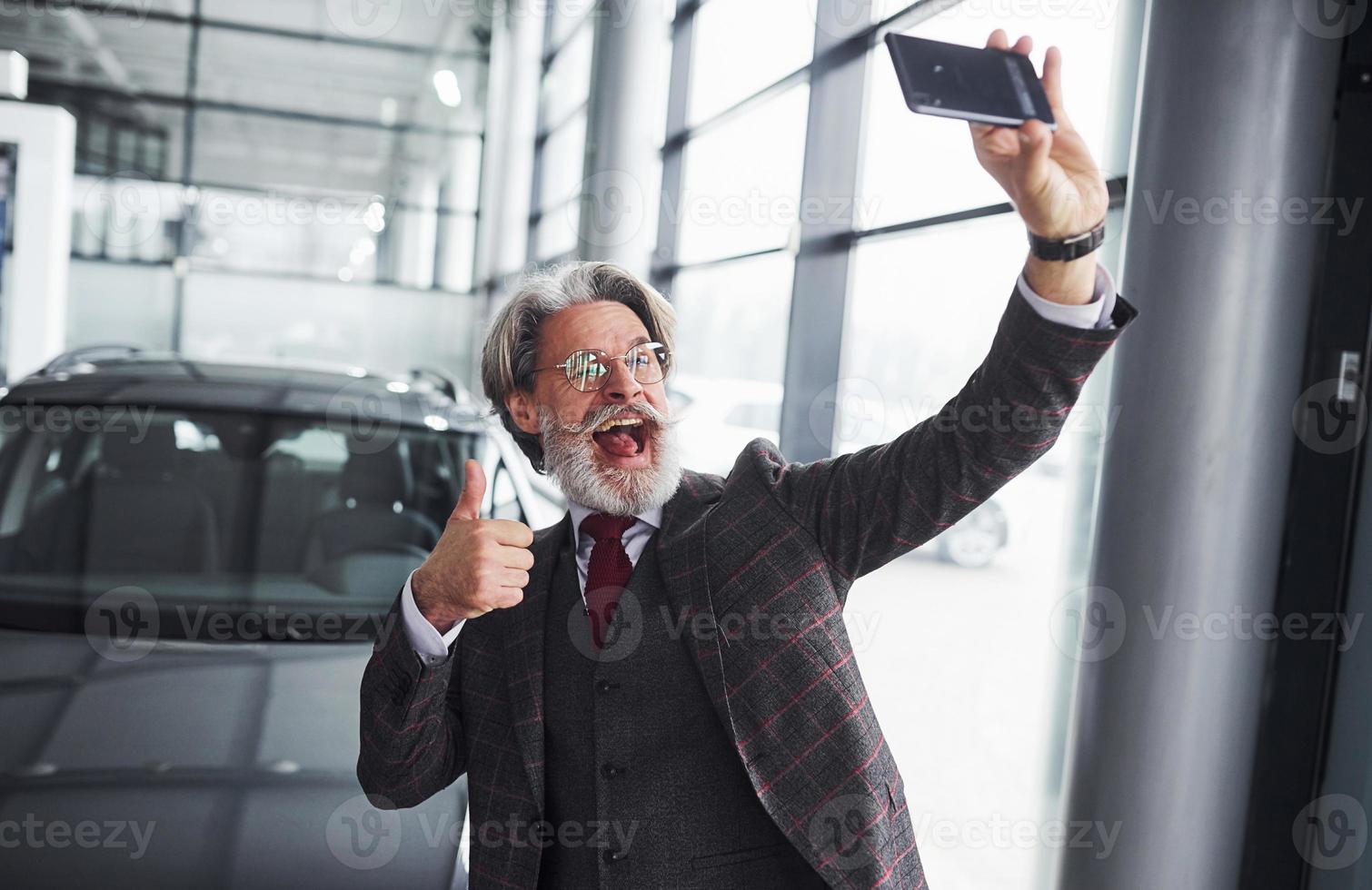 Stilvoller älterer Mann in formeller Kleidung und mit Bart macht Selfie gegen modernes neues Auto. Daumen hoch zeigen foto