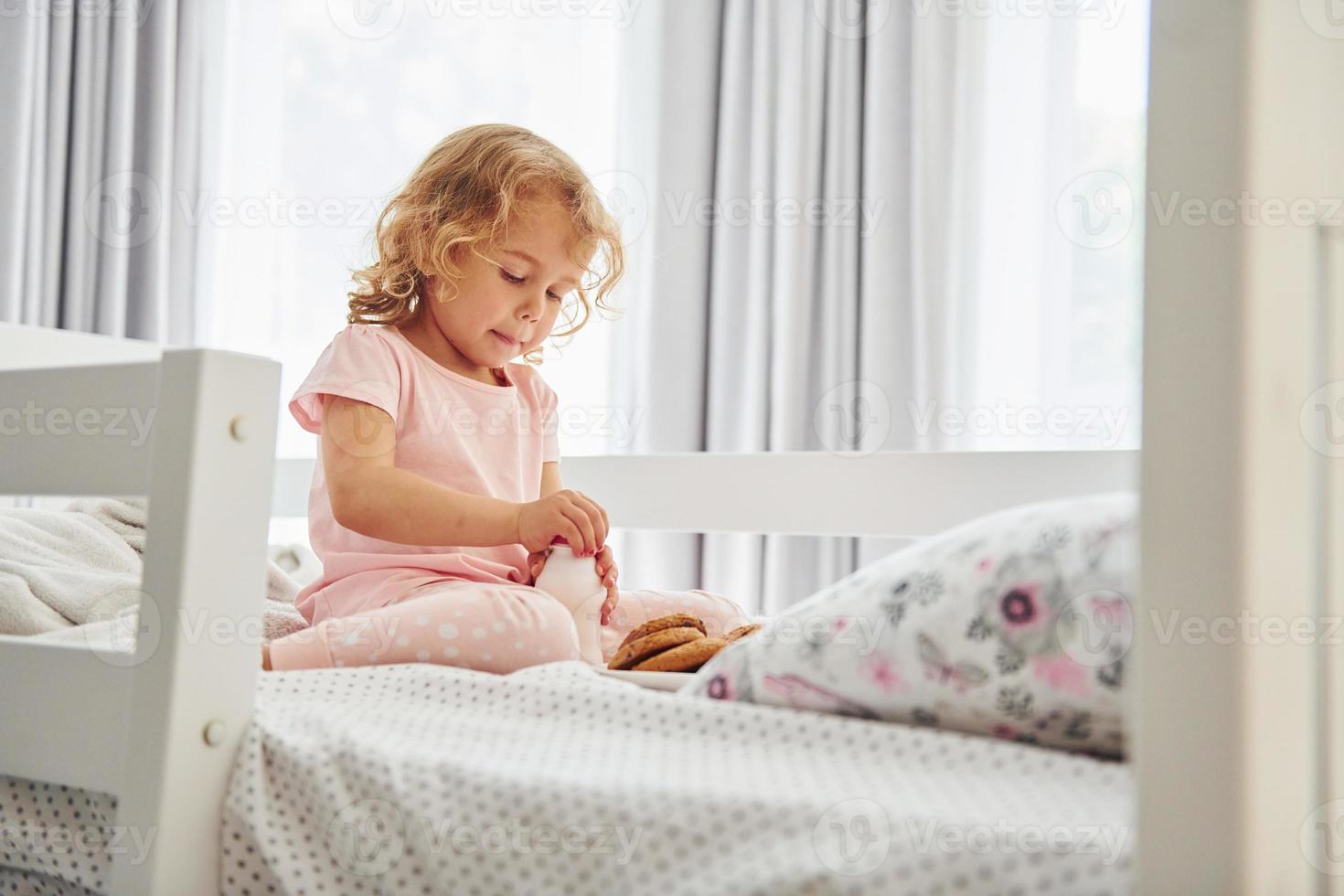 auf dem Bett sitzen, Kekse essen und Milch trinken. süßes kleines mädchen in lässiger kleidung ist tagsüber zu hause foto