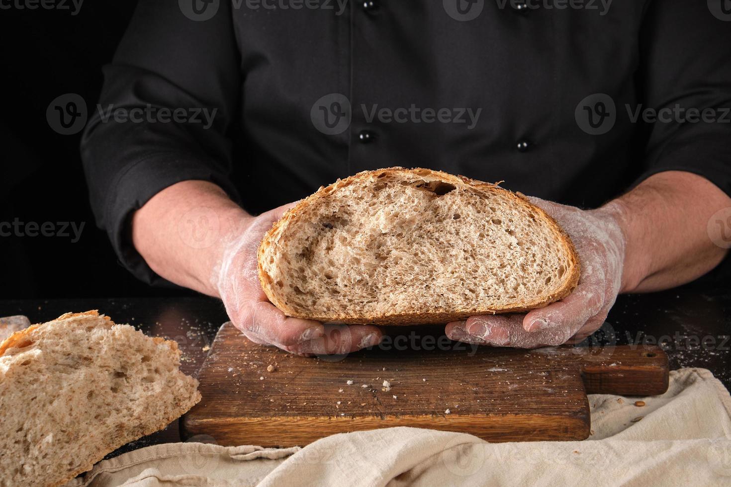 koch in schwarzer uniform hält in seinen händen die hälfte abgebrochenen knusprigen brotes, das aus roggenmehl gebacken wird foto