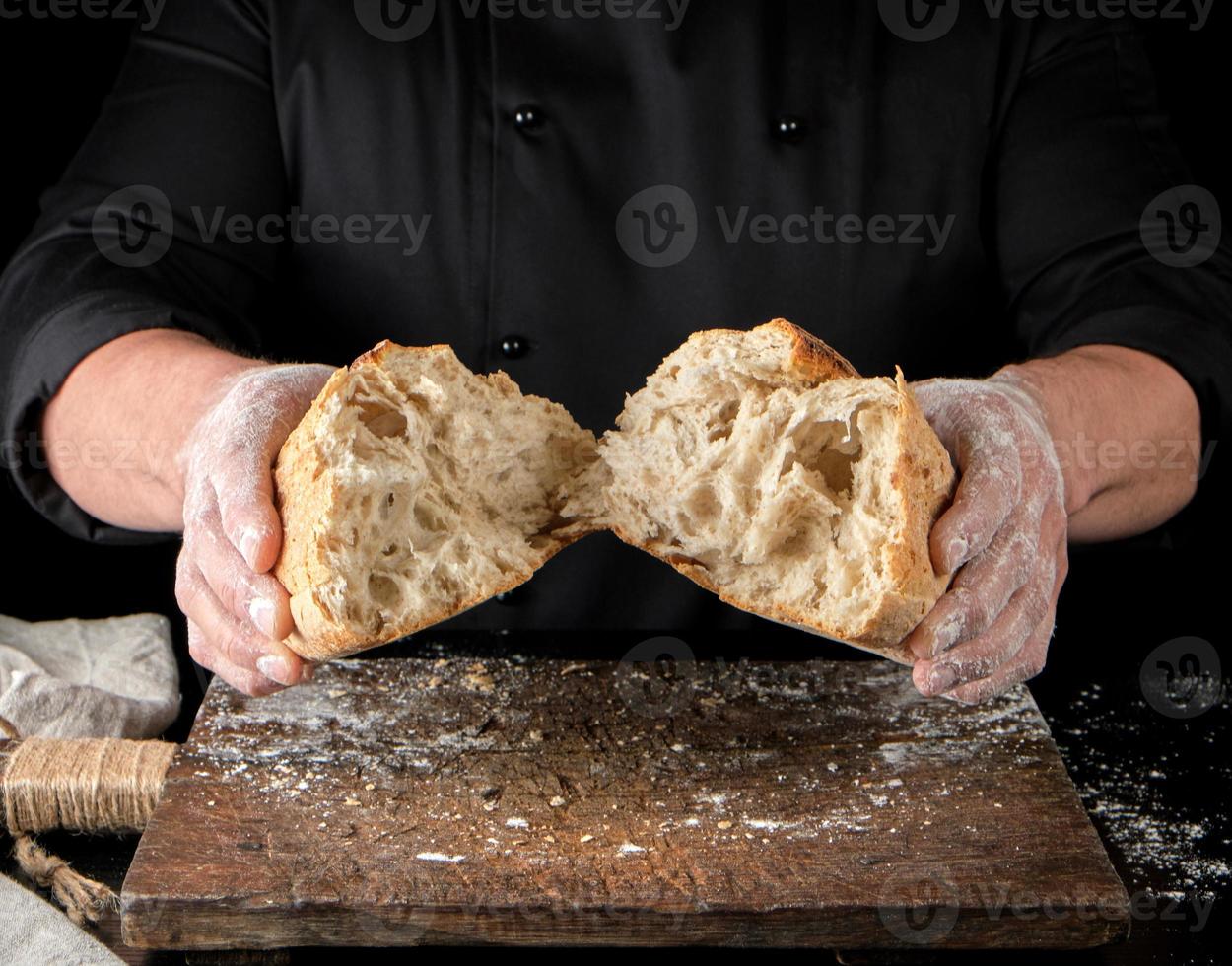 Bäcker in schwarzer Uniform brach einen halben ganzen gebackenen Laib Weißbrot aus Weizenmehl ein foto