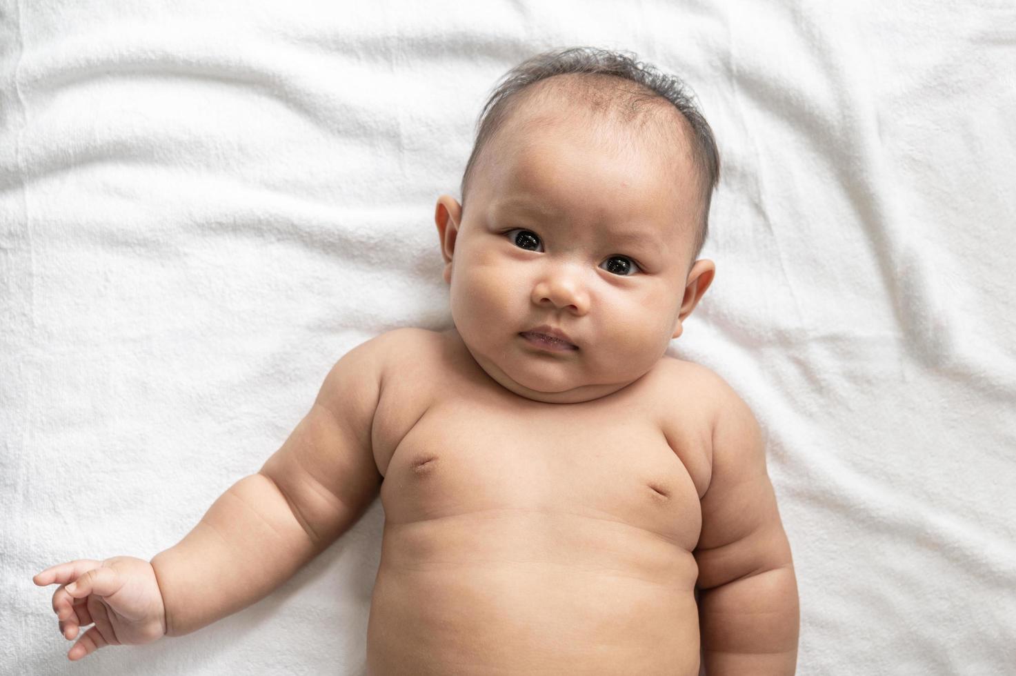 Baby, das Gesicht oben auf einem weißen Bett liegt und Kamera betrachtet foto