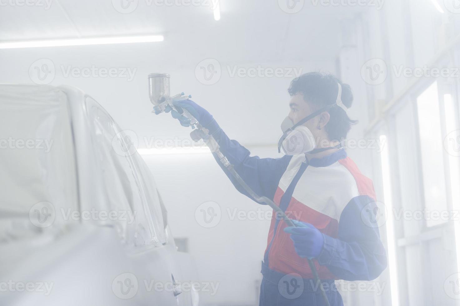 asiatischer automechaniker mit schutzanzug und atemschutzgerät lackiert karosseriestoßstangen im lackierraum und sprüht autos im lackierraum foto