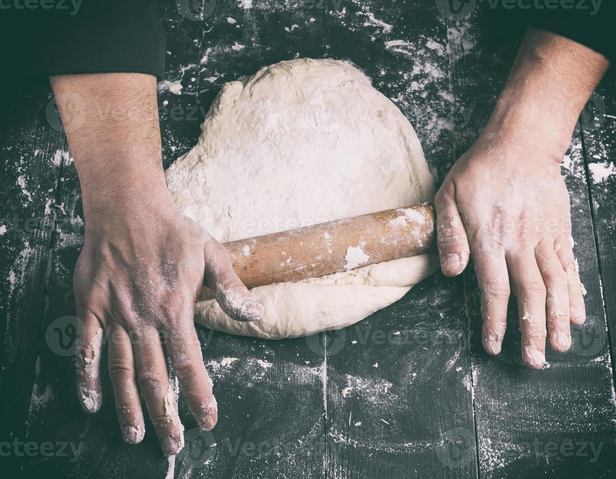 Koch in einer schwarzen Tunika rollt einen Teig für eine runde Pizza foto