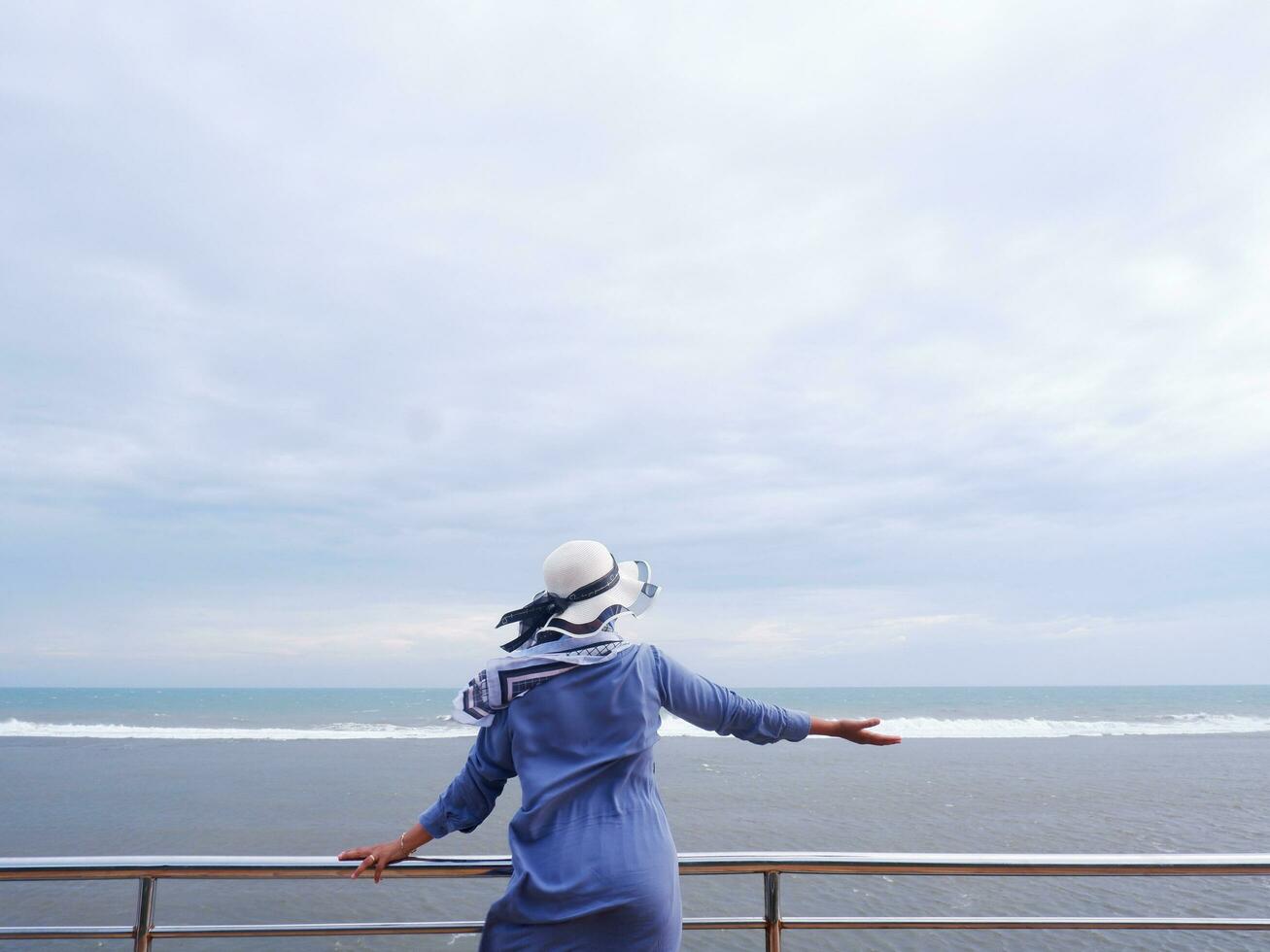 Rücken der Frau mit Hut am tropischen Strand, die den Himmel und das Meer betrachtete, während sie ihre Hände auf der Brücke ausbreitete. Seeblick foto
