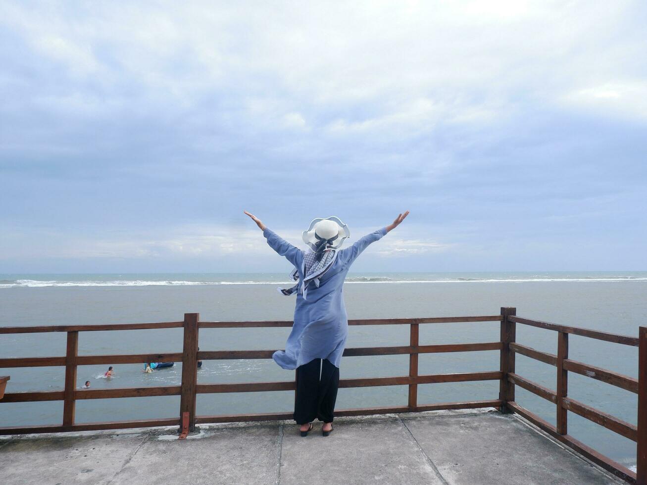 Rücken der Frau mit Hut am tropischen Strand, die den Himmel und das Meer betrachtete, während sie ihre Hände auf der Brücke ausbreitete. Seeblick foto