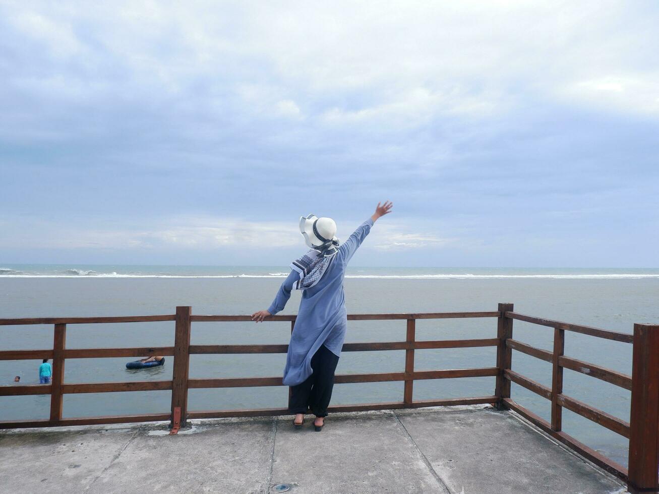 Rücken der Frau mit Hut am tropischen Strand, die den Himmel und das Meer betrachtete, während sie ihre Hände auf der Brücke ausbreitete. Seeblick foto