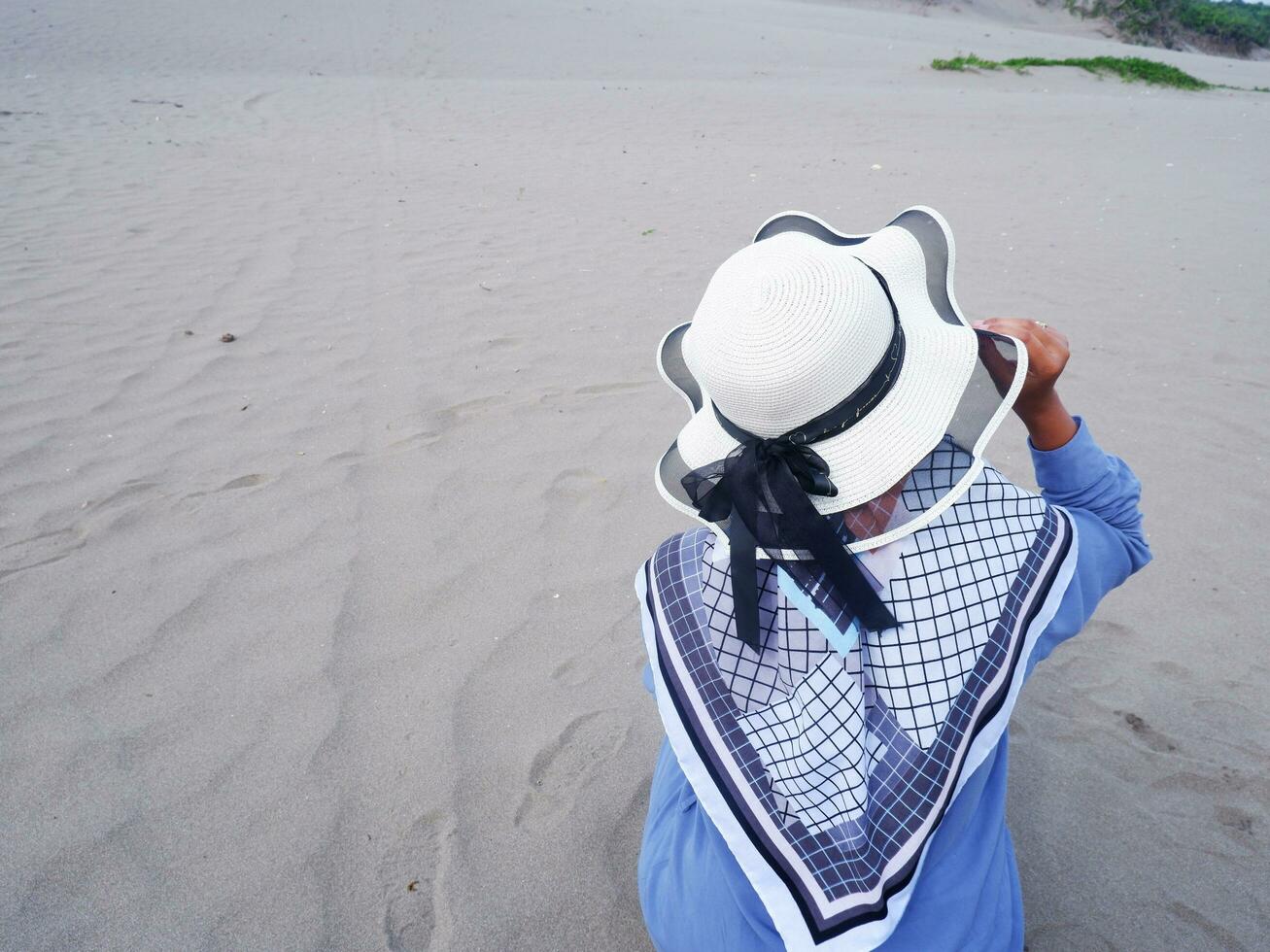 der Rücken der Frau mit Hut am tropischen Strand, die auf dem Strandsand saß. Hintergrund von Strandsand und -himmel. Sandberg foto