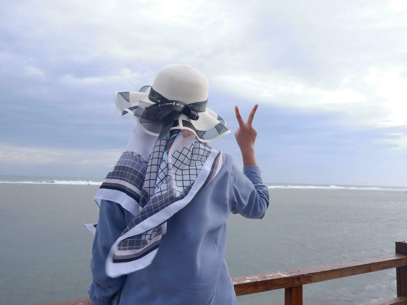 Rückseite der Frau mit Hut am tropischen Strand, die auf den Himmel und das Meer blickt und eine Friedensgeste zeigt. Vertikale. Meereshintergrund foto