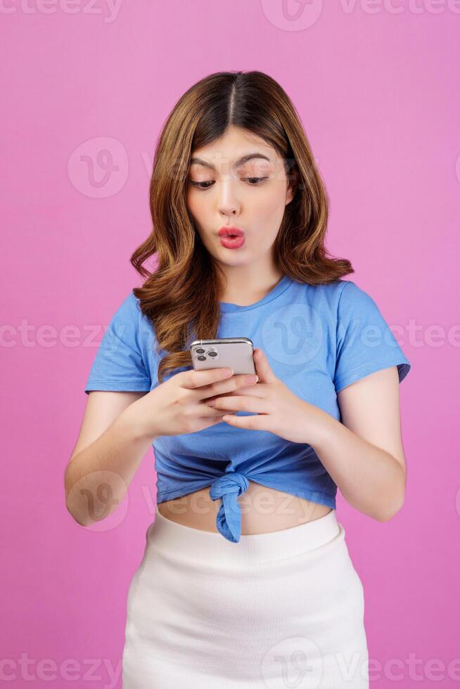 Porträt einer überraschten jungen Frau, die ein Handy in ihren Händen hält, während sie isoliert vor rosa Hintergrund steht foto