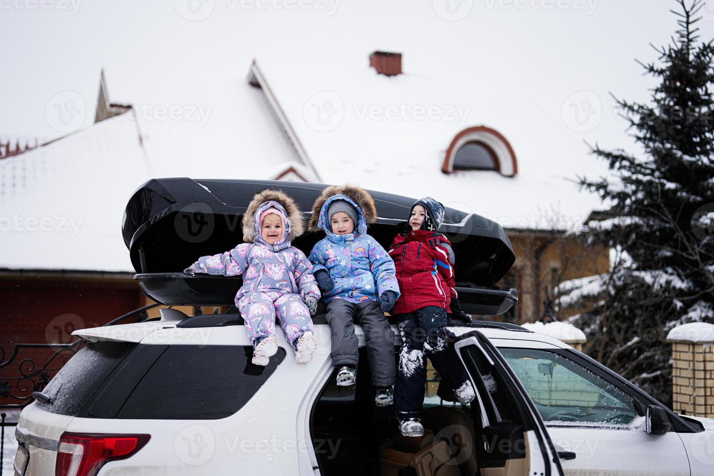 Drei Kinder sitzen im Winter auf einer Autodachbox gegen Haus. foto