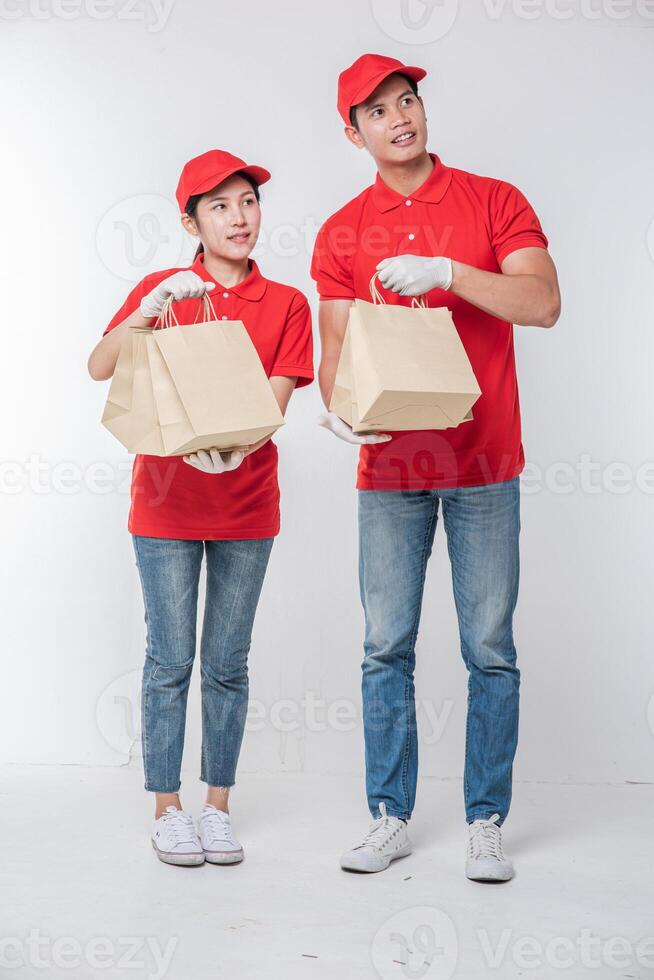 Bild eines glücklichen jungen Zustellers in roter Kappe, leerem T-Shirt, Uniform, stehend mit leerem braunem Kraftpapierpaket, isoliert auf hellgrauem Hintergrund Studio foto